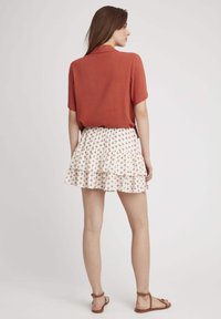 Chemise en rust à manches courtes avec un col, associée à une jupe blanche à volants ornée de motifs floraux rouges. Des sandales plates marron complètent la tenue.