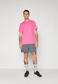 Rosa treningst-skjorte, grå shorts, svarte joggesko og grå sokker med logo; modellen står oppreist og viser tatoveringer på armer og bein.