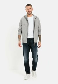 Grå zip-hoodie med hætte og brune accenter, båret over en hvid T-shirt. Parret med mørkeblå jeans og hvide sneakers.