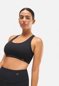 Sujetador deportivo negro con tirantes anchos, escote redondeado y textura suave. Presenta un pequeño logo impreso en la banda.