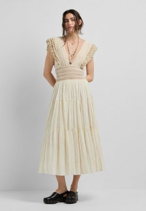 Frau trägt ein ärmelloses, cremefarbenes Kleid mit strukturiertem Oberteil und gestuftem Rock, geschichtete Halsketten und schwarze Holzschuhe, steht vor einer schlichten Wand.