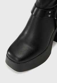Bottine en cuir noir avec une plateforme épaisse et un talon carré. Dotée d'un bout arrondi et d'une sangle décorative avec des accents métalliques.