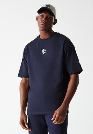 Mørkeblå bomulds t-shirt med korte ærmer, rund halsudskæring og et hvidt logo på brystet. Sammen med en beige kasket med et mørkt logo.