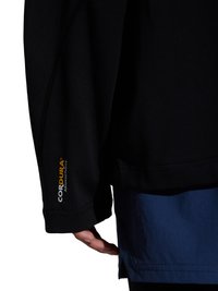 Zbliżenie na ramię osoby noszącej czarny rękaw z tkaniny Cordura oraz niebieski strój pod spodem, dłoń częściowo widoczna pod rękawem.