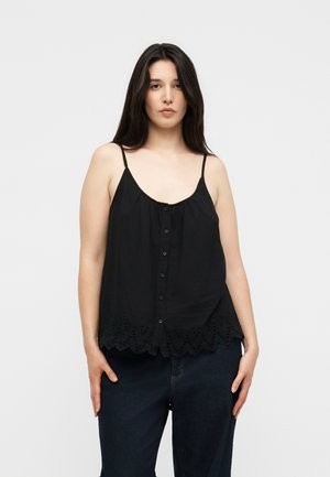 Jonge vrouw met lang donker haar draagt een zwarte mouwloze top met knopen en een kanten zoom en donkerblauwe jeans, staand tegen een witte achtergrond.