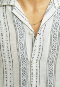 Chemise légère à manches courtes en lin de couleur crème avec des motifs géométriques bleus verticaux, un col à encoche et un devant à boutons.