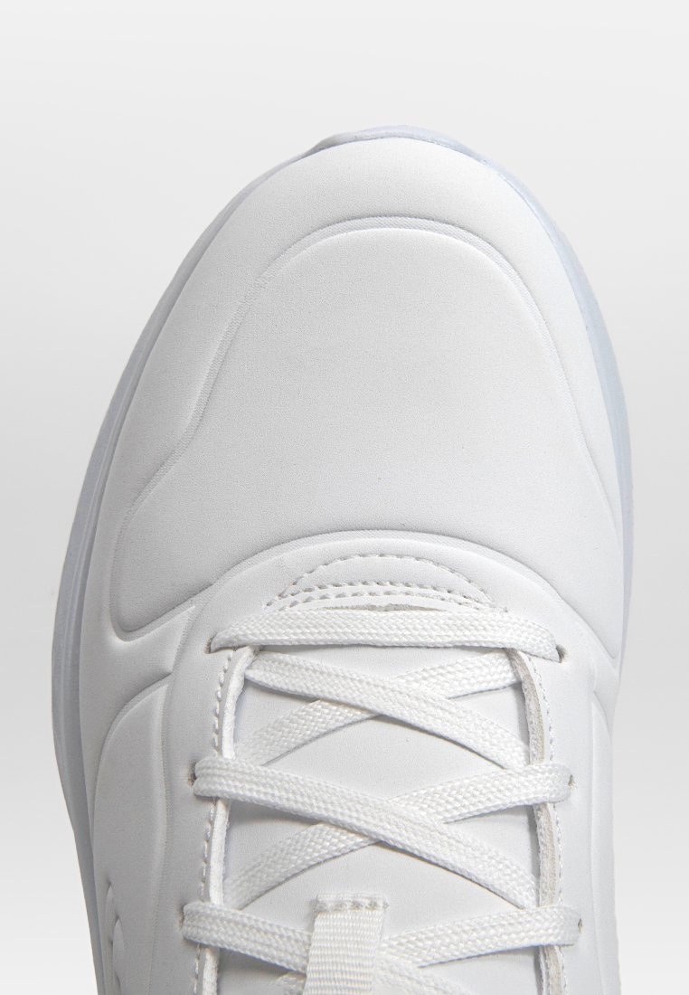 Zapatilla deportiva blanca con una parte superior sintética suave, diseño de cordones, acentos texturizados y una suela semitranslúcida.