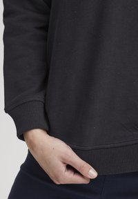 Felpa nera con polsini e orlo a coste, che mostra una texture liscia. La mano poggia sull'orlo, indicando una vestibilità e un design rilassati.