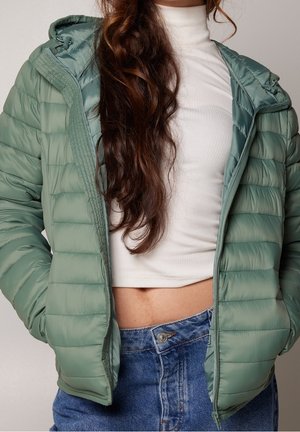 Persoon met lang bruin haar die een open lichtgroene donzen jas, een witte cropped top en blauwe high-waisted jeans draagt met de handen in de zakken.