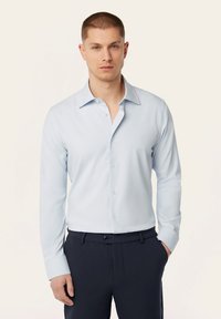  Lichtblauw overhemd met lange mouwen en een puntkraag, met een knoopsluiting en een gladde textuur. Gedeeltelijk gedragen met donkere broek en handen in de zakken.