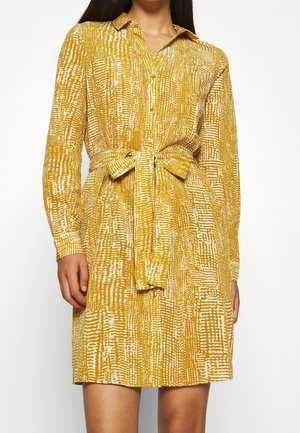 Sukienka koszulowa w kolorze musztardowym z białym abstrakcyjnym wzorem; posiada kołnierzyk, długie rękawy i dopasowany pasek w talii.