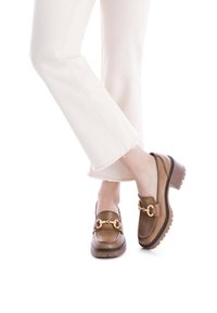 Mocasines de cuero marrón con un tacón grueso, detalle de cadena dorada y suela texturizada. Usados con pantalones blancos desgastados.