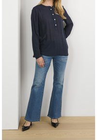 Blusa a maniche lunghe blu navy con apertura frontale a bottoni, abbinata a jeans a zampa d'elefante azzurri e décolleté nere a punta. Tessuto morbido con una leggera lucentezza.