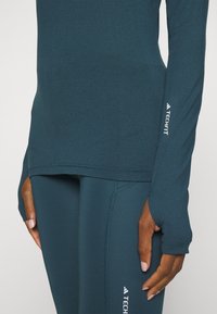 Teal långärmad träningsblus och matchande leggings har fukttransporterande tyg, åtsittande passform och tumhål på ärmarna. TECHFIT-logotyp synlig.