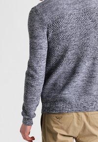 Szary sweter z dzianiny z teksturowanym wzorem, z prążkowanym obszyciem na dole i mankietach. Tył ozdobiony wzorem przypominającym jodełkę.