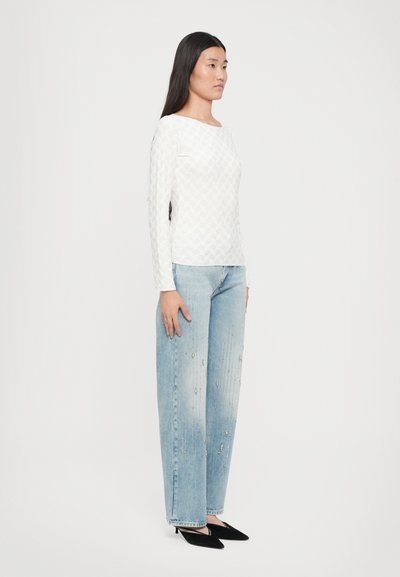 Hvit langermet topp med et teksturert geometrisk mønster, kombinert med lyseblå vide jeans med metalliske detaljer. Svarte spissede sko.