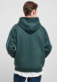 Sudadera con capucha en verde azulado oscuro, hecha de un suave forro polar. Presenta un bolsillo tipo canguro y puños y dobladillo acanalados. Vista trasera, usada sobre una camiseta blanca.