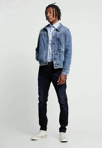 Chaqueta de mezclilla en azul claro, abotonada con bolsillos en el pecho; llevada sobre una camisa azul clara, combinada con jeans azul oscuro y zapatillas blancas.