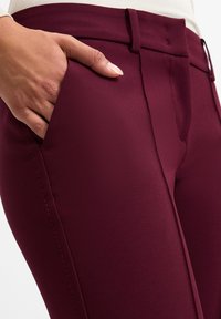 Bordeaux kleurige op maat gemaakte broek van een soepele, rekbare stof. Heeft een slim fit, zijzakken en zichtbare stiksels langs de naden.