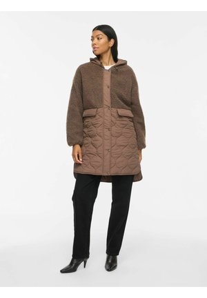 Vrouw draagt een bruine hooded jas met doorgestikte onderste helft en fleece bovenste helft, zwarte broek en zwarte laarzen met hak, staat met ontspannen handen.