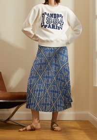 Nainen, joka on pukeutunut valkoiseen collegepaitaan, jossa lukee "Sandro 1984 Pariisi", ja siniseen kuvioituun midi-hameeseen, seisoo sisätiloissa ruskean tuolin vieressä.