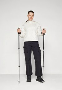 Chaqueta blanca con cremallera, pantalones cargo negros y calzado negro. Sosteniendo dos bastones de trekking negros. Fondo simple.