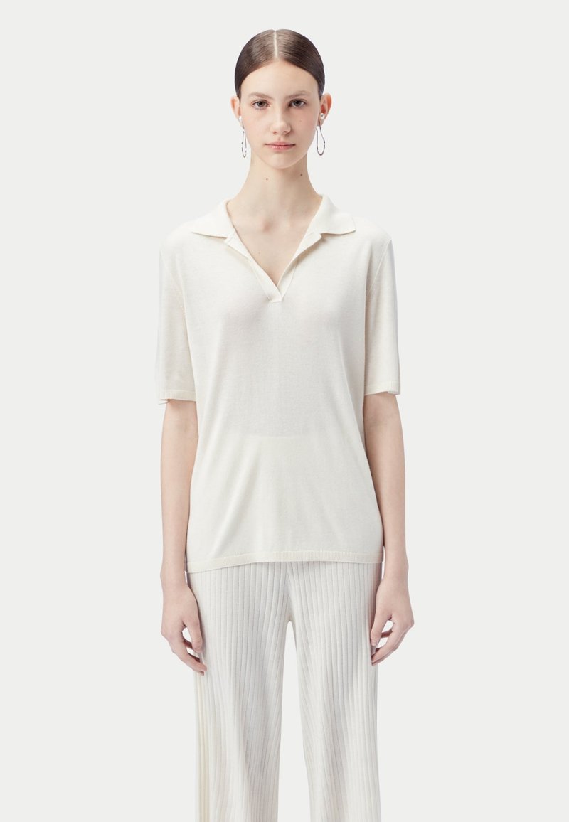 Ung kvinna med bakåtslickat hår bär silverfärgade creoler, en krämfärgad kortärmad skjorta med krage och krämfärgade ribbade byxor, stående mot en vit bakgrund.