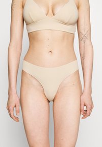 Beige, varrat nélküli melltartó és tanga szett, puha, sima anyagból készült. A melltartó V-nyakú dizájnnal rendelkezik, a tanga pedig minimalista szabású.