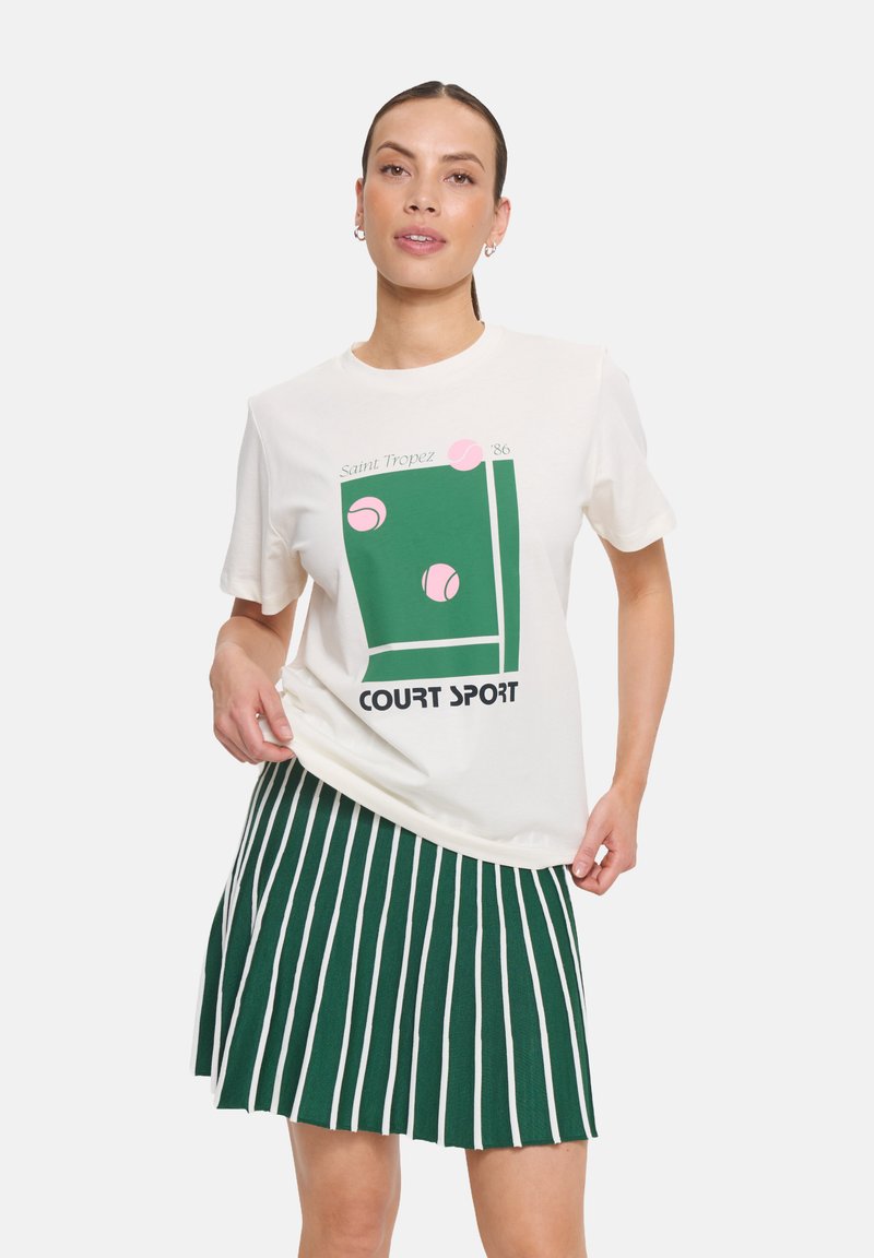 T-shirt crème avec un graphisme vert comprenant des balles de tennis roses et le texte "Saint Tropez '86", associé à une jupe plissée rayée verte.