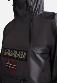 Grå udendørsjakke med en lynlåslomme, prydet med et logo patch og et norsk flag emblem. Materialet har en glat, blank overflade.