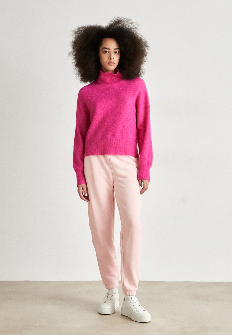 Un pull col roulé fuchsia en tricot associé à un pantalon de survêtement rose clair et des baskets blanches. Texture douce, coupe décontractée, poignets élastiques sur le pantalon.