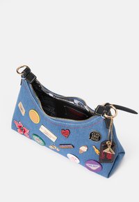 Denimaxelväska med flerfärgade lappar och nålar, dragkedjestängning, svart läderrem och röda sömdetaljer på en blå tygbakgrund.