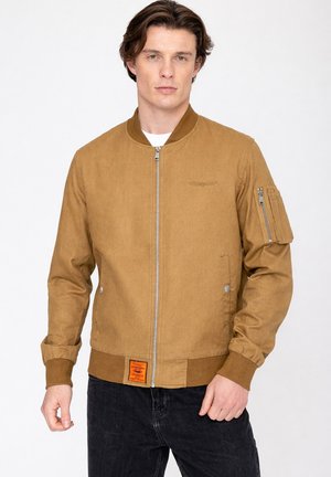 Junger Mann trägt eine braune Bomberjacke mit Reißverschluss, gerippten Bündchen und Saum, Reißverschlusstasche am linken Ärmel und schwarze Jeans.