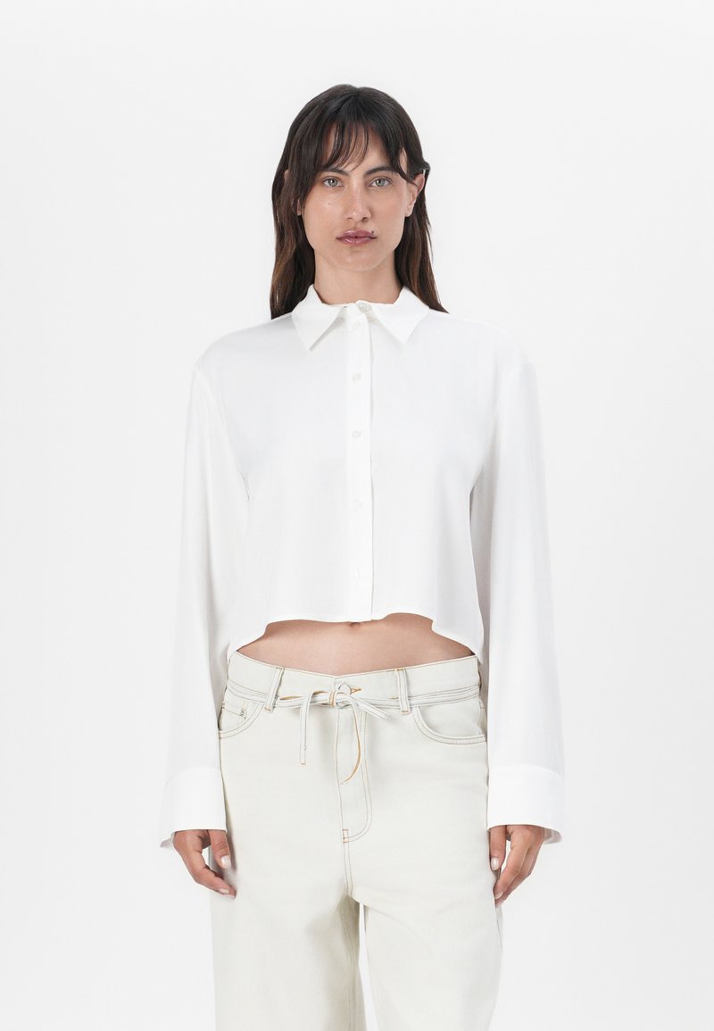 White cropped button-up shirt with a collar and long sleeves. Smooth texture, featuring a straight hem and subtle button detailing.