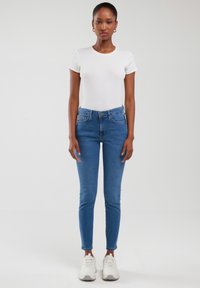 Naine seisab, kandes valget kehakuju rõhutavat t-särki, siniseid kõrge vöökohaga kitsaid teksapükse, valgeid tossusid ja kuldseid rõngakujulisi kõrvarõngaid ühtlasel taustal.