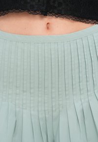 Φιστικί πλισέ φούστα με δομημένη μέση. Διαθέτει οριζόντιες και κάθετες πτυχώσεις, κατασκευασμένη από ελαφρύ ύφασμα με λεία υφή.