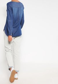 Camicia di jeans blu scuro, con scollo tondo, maniche lunghe e orlo curvo. Abbinata a pantaloni grigi chiari e sneaker bianche.