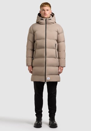 Beige gewatteerde jas met een capuchon, met zwarte ritsdetails en twee zijzakken; gedragen over zwarte broeken en laarzen; gladde textuur.
