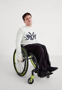 Bílá mikina s modrým plamenným motivem, černé volné kalhoty a černé invalidní vozíku se zelenými akcenty a velkými černými koly.