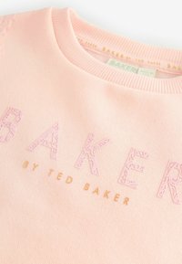 Sweatshirt rose clair avec col côtelé, comportant le mot "BAKER" en lettres texturées rose et or. Texture de tissu douce et lisse.