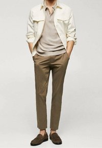 Hombre con chaqueta beige sobre una camiseta polo beige clara, pantalones cortos marrones y mocasines marrones, de pie con las manos en los bolsillos.