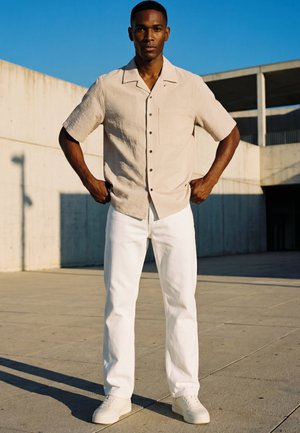 Un homme se tient à l'extérieur sur une surface en béton, portant une chemise beige à manches courtes boutonnée, un pantalon blanc et des baskets blanches, les mains sur les hanches.