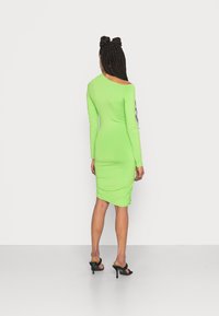 Vestido verde de manga longa com um design assimétrico, silhueta ajustada, detalhes franzidos nas costas e sandálias de salto pretas.
