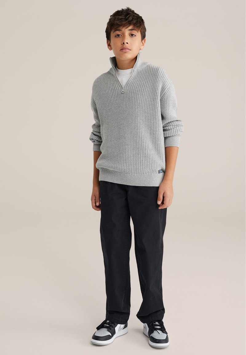 Svetlý sivý rebrovaný pletený sveter s golierom na zips, skombinovaný s čiernymi nohavicami a čierno-sivými teniskami. Jednoduchý, pohodlný dizajn bez viditeľných vzorov.