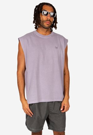 Uomo con dreadlocks e occhiali da sole che indossa una maglietta lavanda senza maniche e pantaloncini grigio scuro, in piedi su uno sfondo bianco.