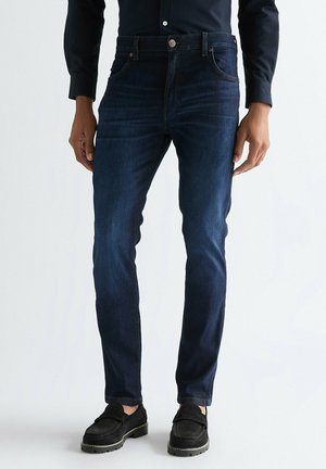 Mężczyzna ubrany w ciemnoniebieskie dopasowane jeansy, czarną koszulę na guziki oraz czarne mokasyny, stojący na jednolitym białym tle.