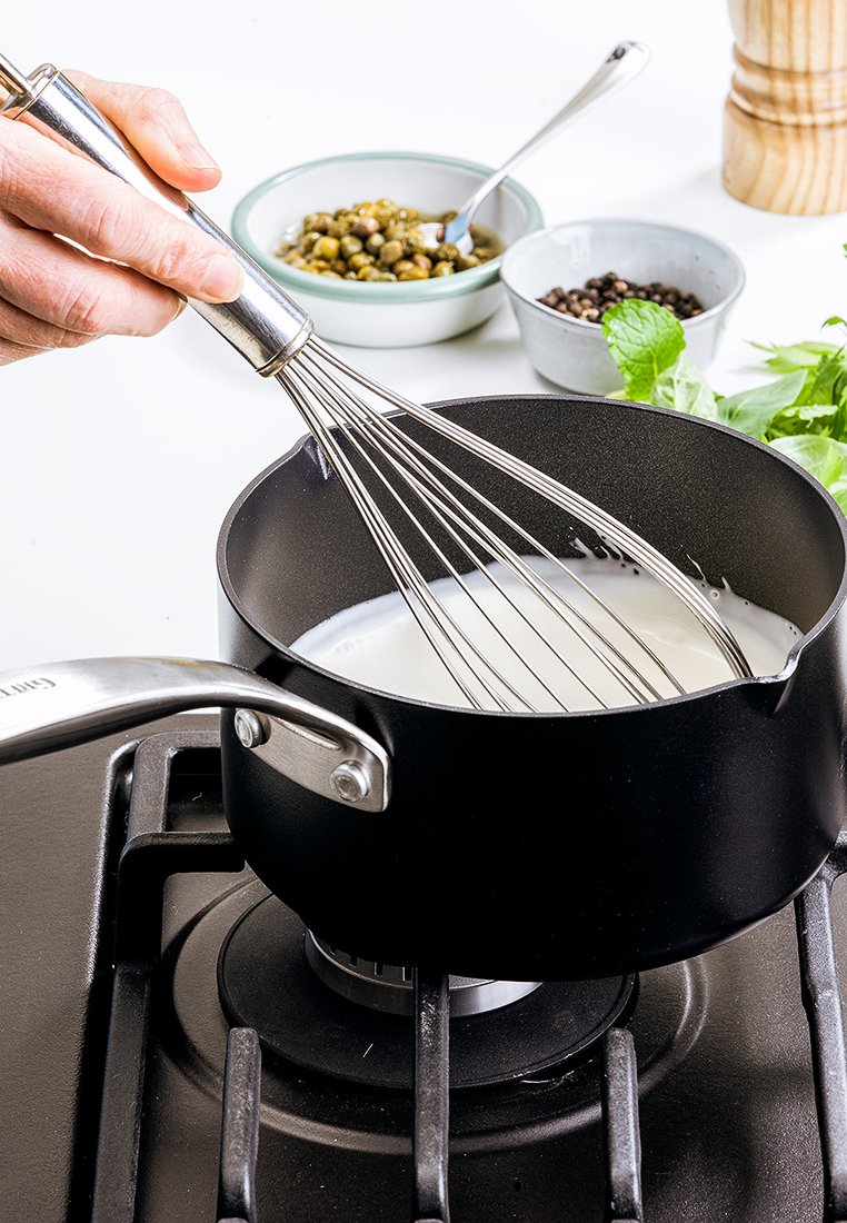 Czarny garnek na kuchence gazowej, wypełniony białym płynem, używana metalowa trzepaczka. Wokół znajdują się miseczki z ziołami i przyprawami.