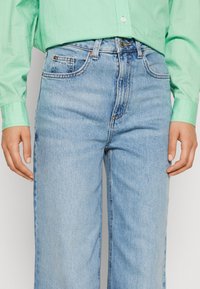 Jeans de tiro alto en azul claro con pierna recta, que cuentan con cinco bolsillos y un cierre de botón, combinados con una camisa abotonada en verde claro.