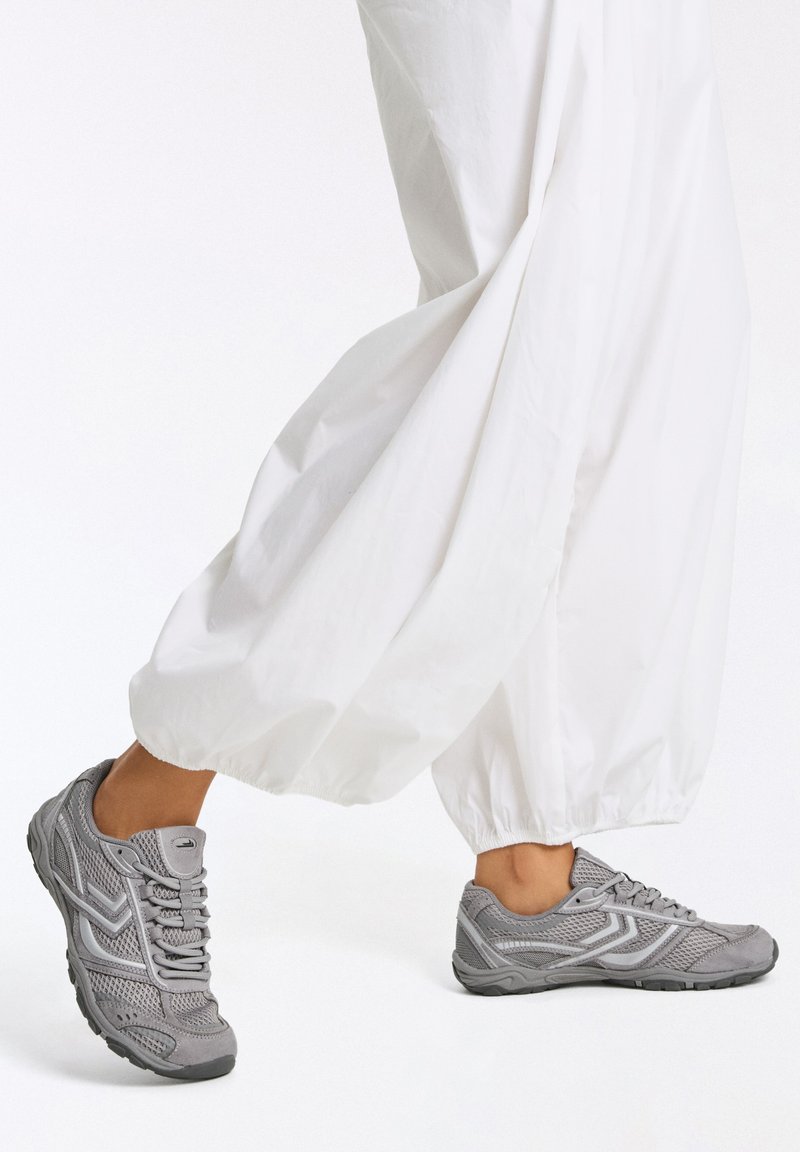 Personne portant un pantalon blanc ample et des chaussures de sport grises marchant sur un fond blanc uni.