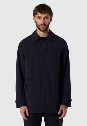 Uomo con capelli castani corti e barba, che indossa un cappotto blu scuro con bottoni e tasche frontali su uno sfondo semplice.